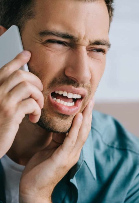young man talking by smartphone and looking away while suffering from tooth pain at home